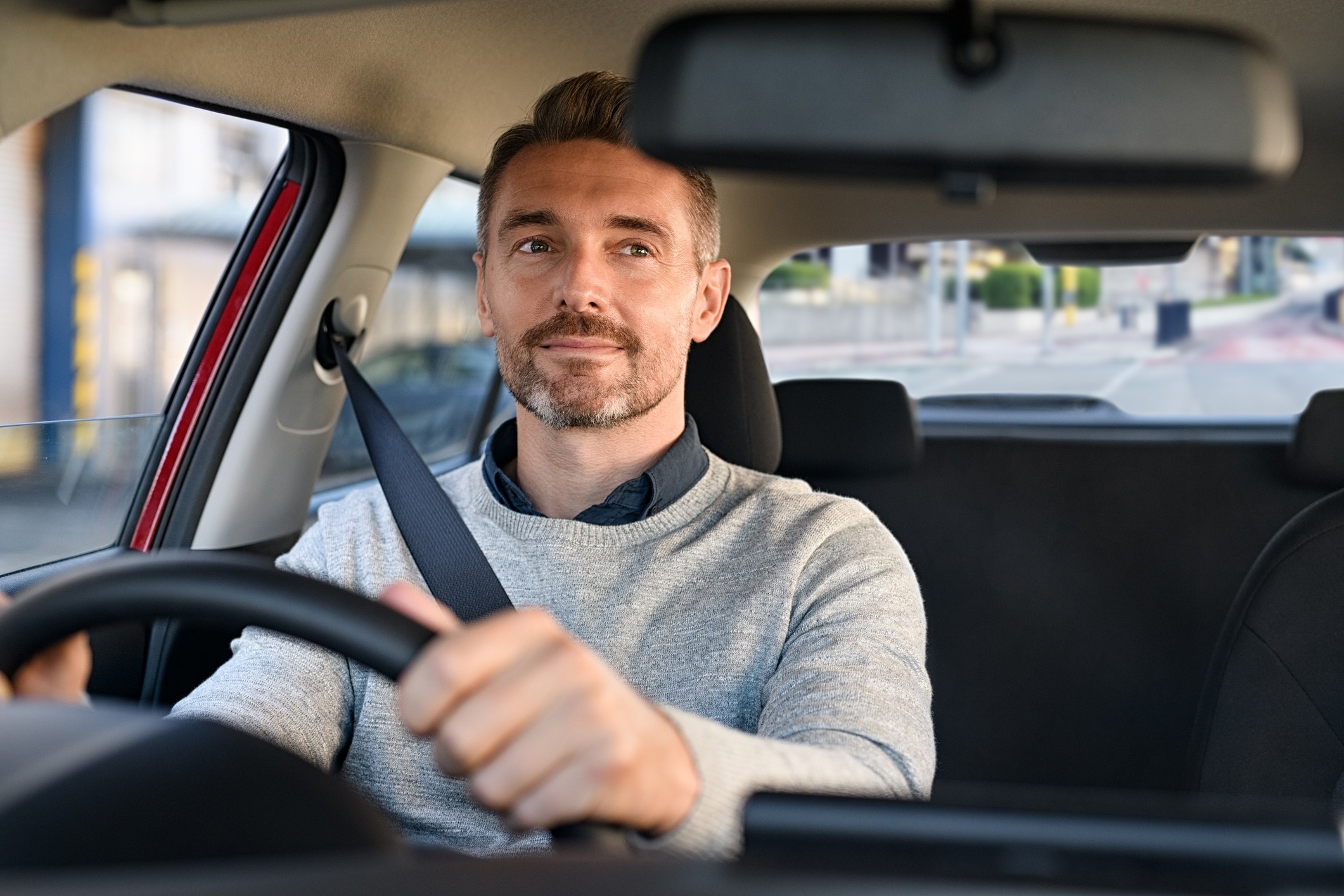 A man driving a car. He is wearing a grey sweater and is looking at the camera with a slight smile.